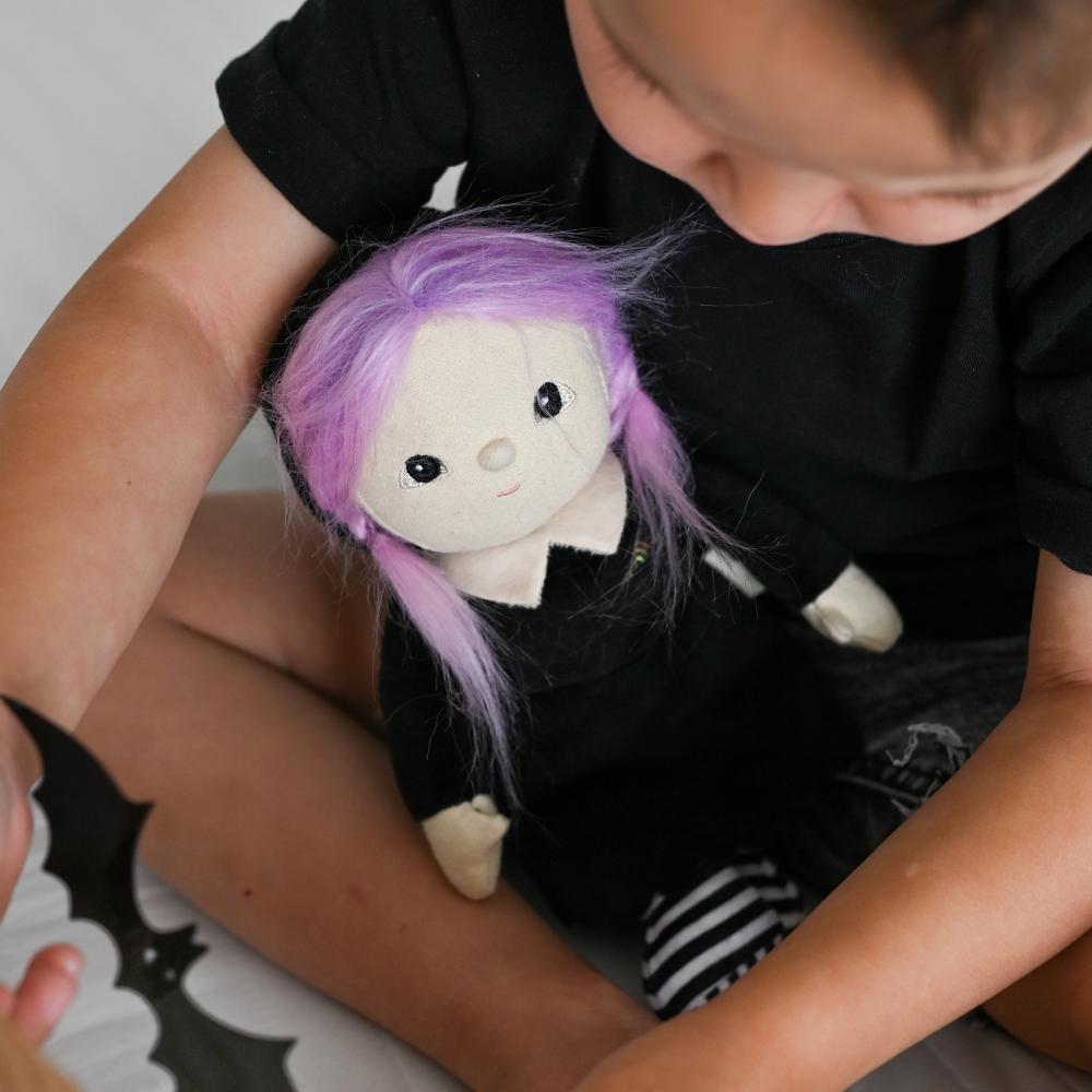 A child in a black shirt holds a small plush doll with purple hair, a white face, and a black outfit with white striped legs.