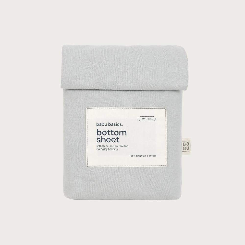 A light gray cotton bottom sheet by babu basics, neatly folded and packaged in a matching fabric sleeve with a white label, set against a solid off-white background.