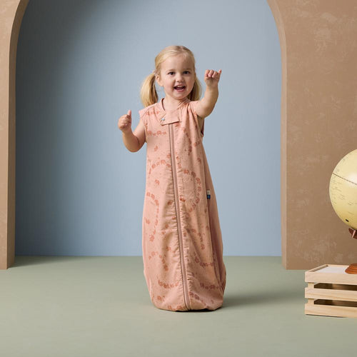 Child in a pink sleep sack standing in a room with a blue wall and wooden floor.
