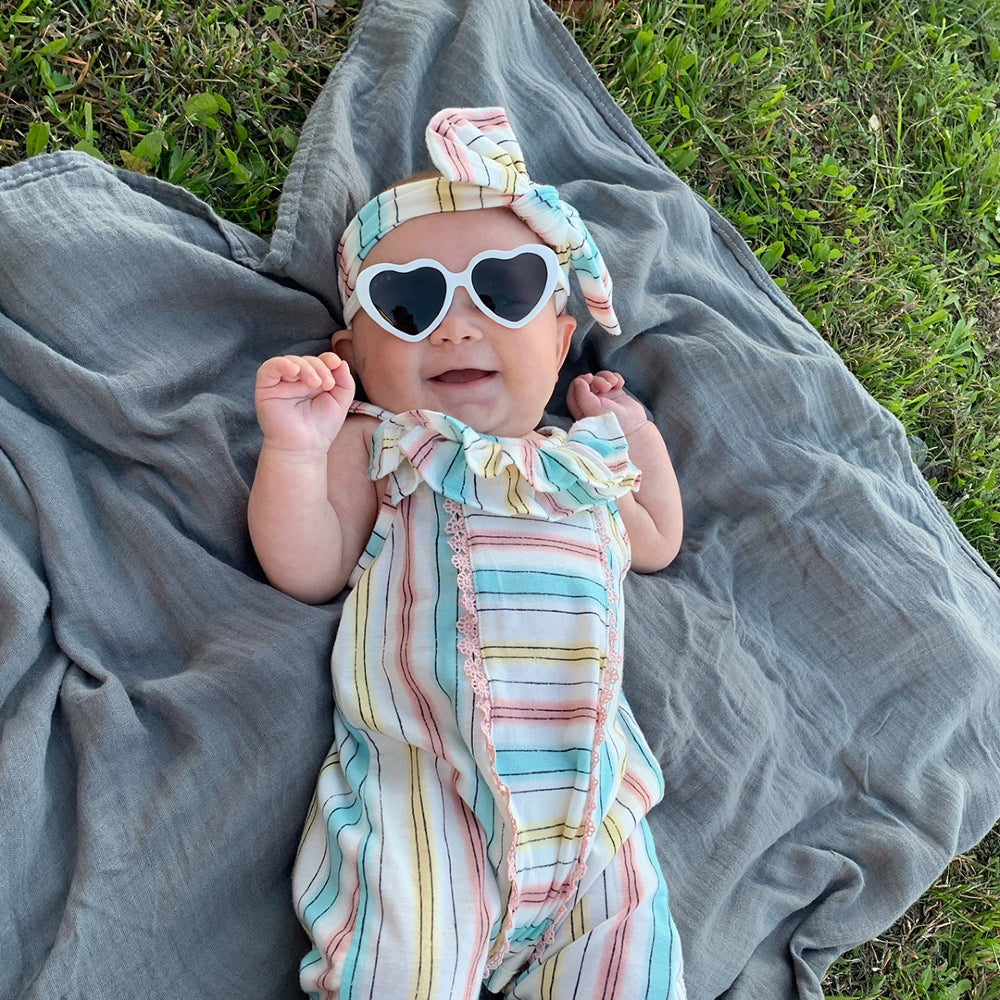 Baby in a striped outfit and sunglasses lying on a gray blanket outdoors.
