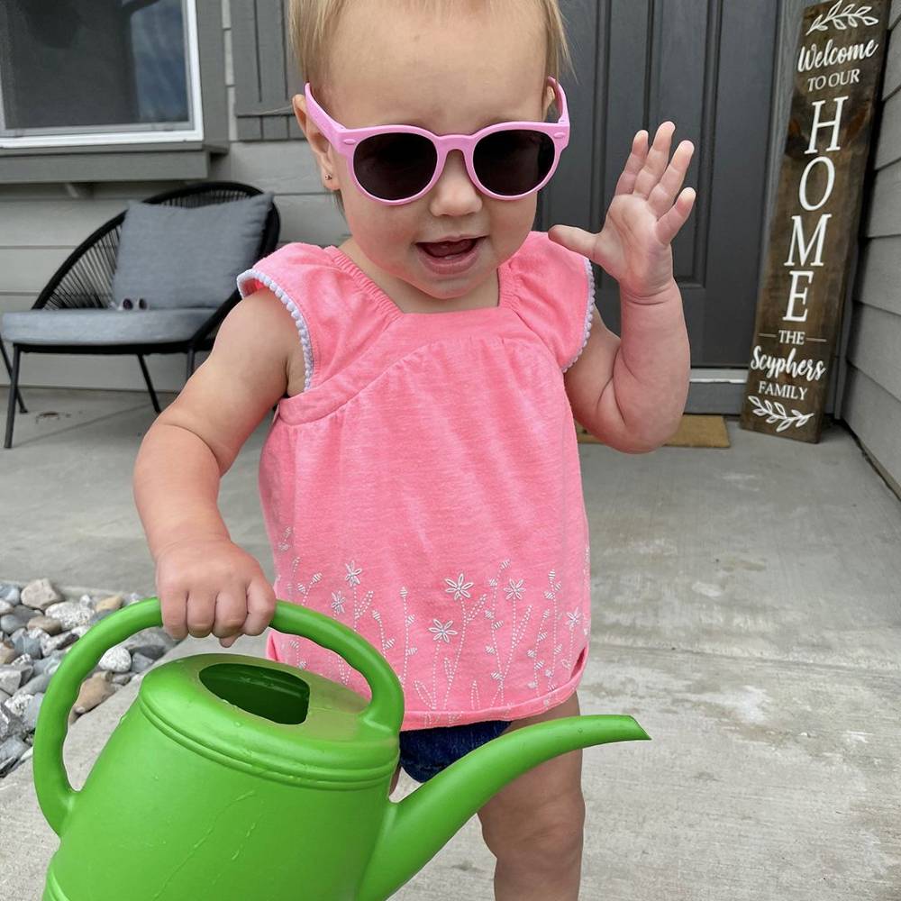 Child wearing pink sunglasses and a pink shirt, holding a green watering can on a porch.