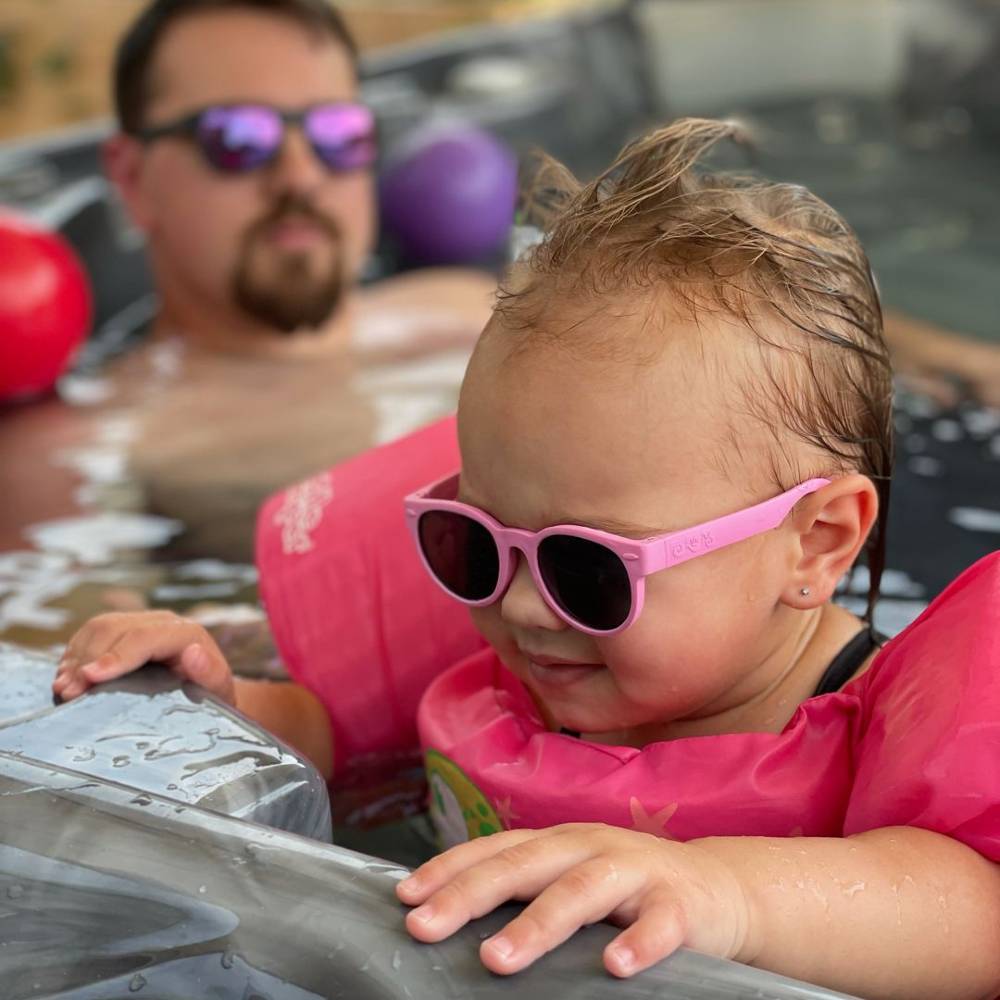 Child wearing pink sunglasses and a life jacket in a pool with a person in the background.