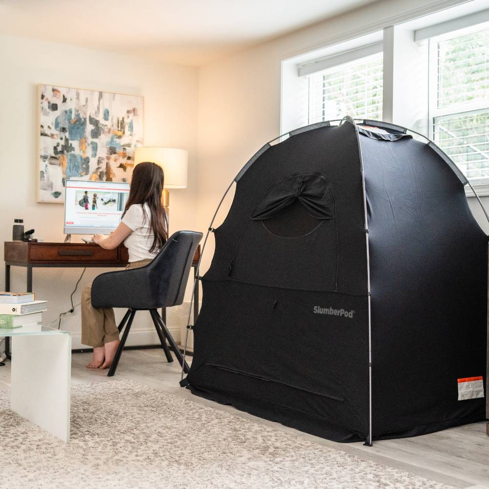 A black Slumberpod pop-up tent with a window and zippered door is set up in a home office environment next to a desk where a woman is working on a computer.
