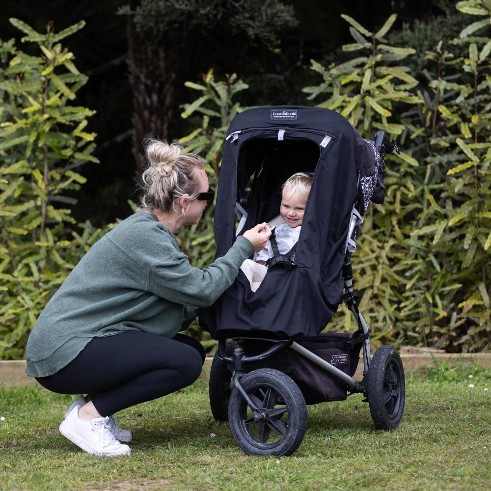 Woman adjusting a stroller with Snoozeshade blackout cover with a child inside in a garden setting.