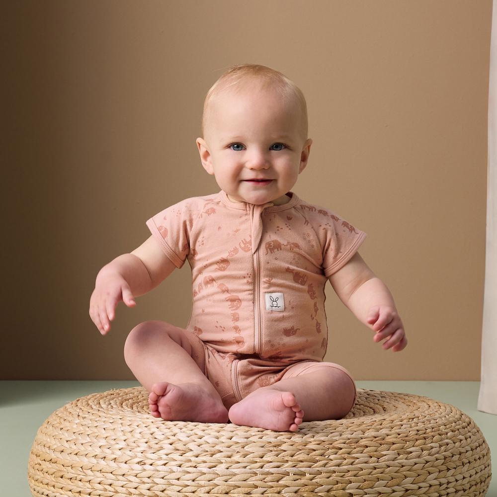 Baby sitting on a woven stool wearing a pink onesie against a beige background