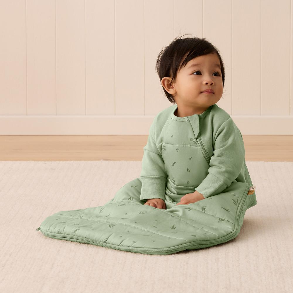 Child wearing a green baby sleep sack sitting on a light wooden floor.