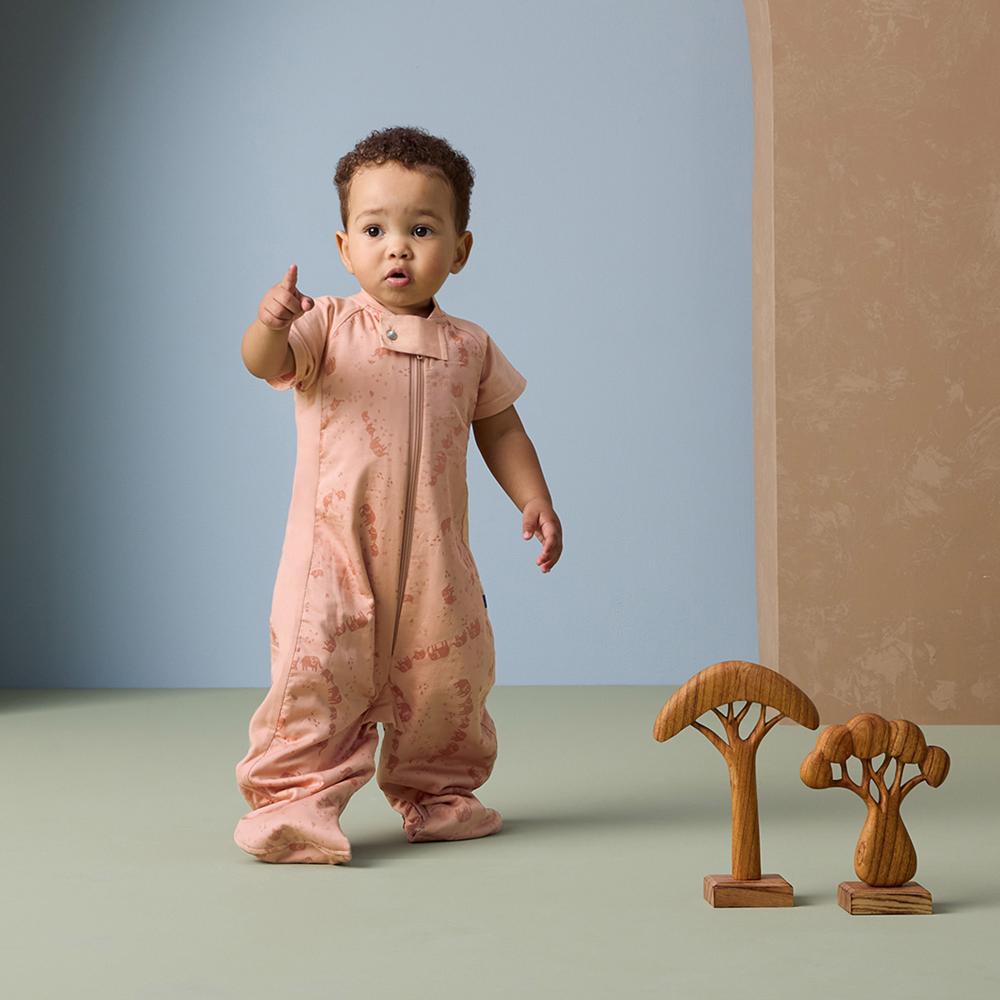Baby in a pink onesie standing next to wooden tree toys on a light green surface with a blue wall background.
