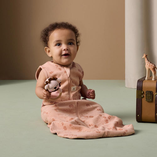 Baby in a pink elephant pint sleeping bag with a toy, sitting next to a suitcase and giraffe toy on a beige background