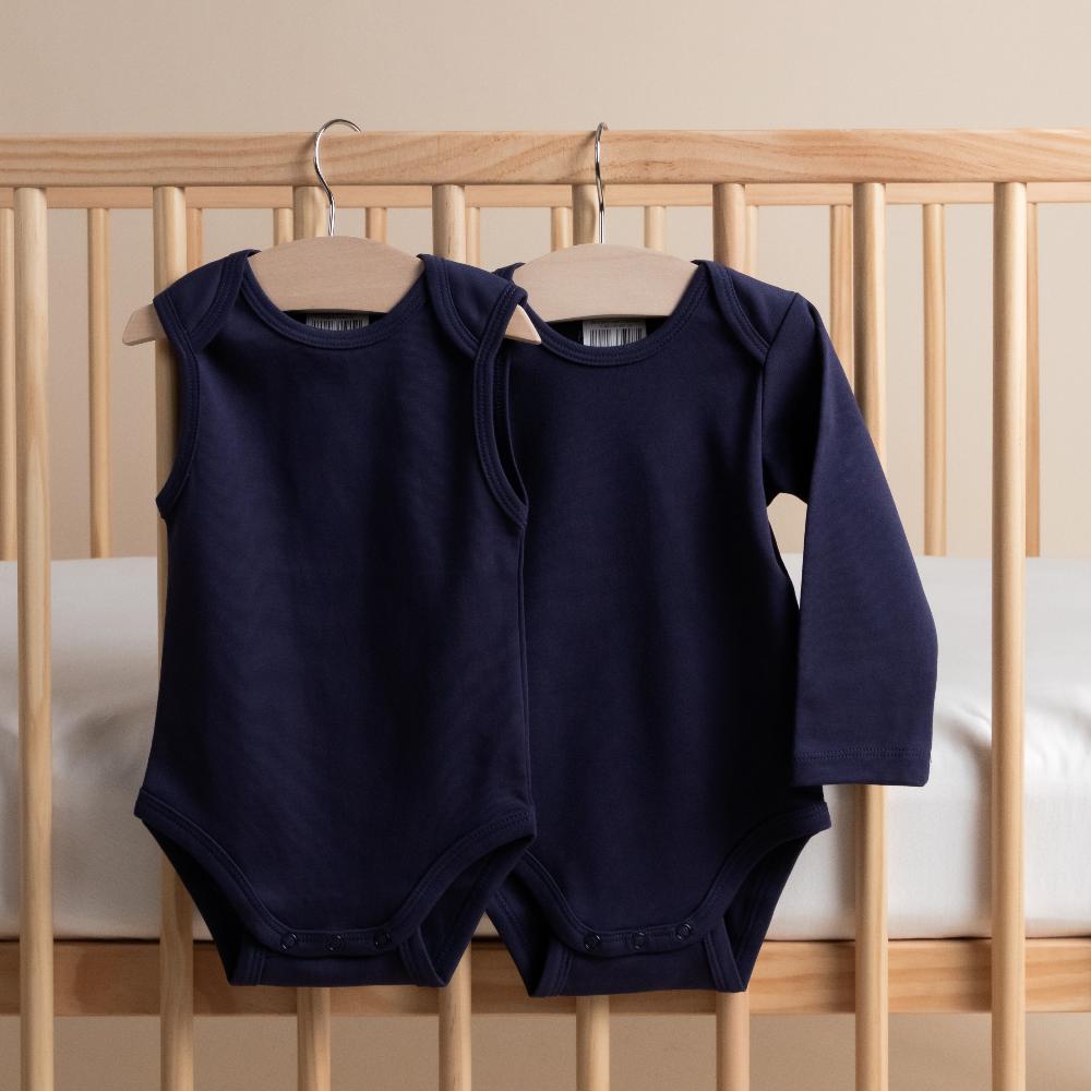 Two navy blue organic cotton baby bodysuits, one singletsuit and one long sleeve on hangers against a wooden crib background.