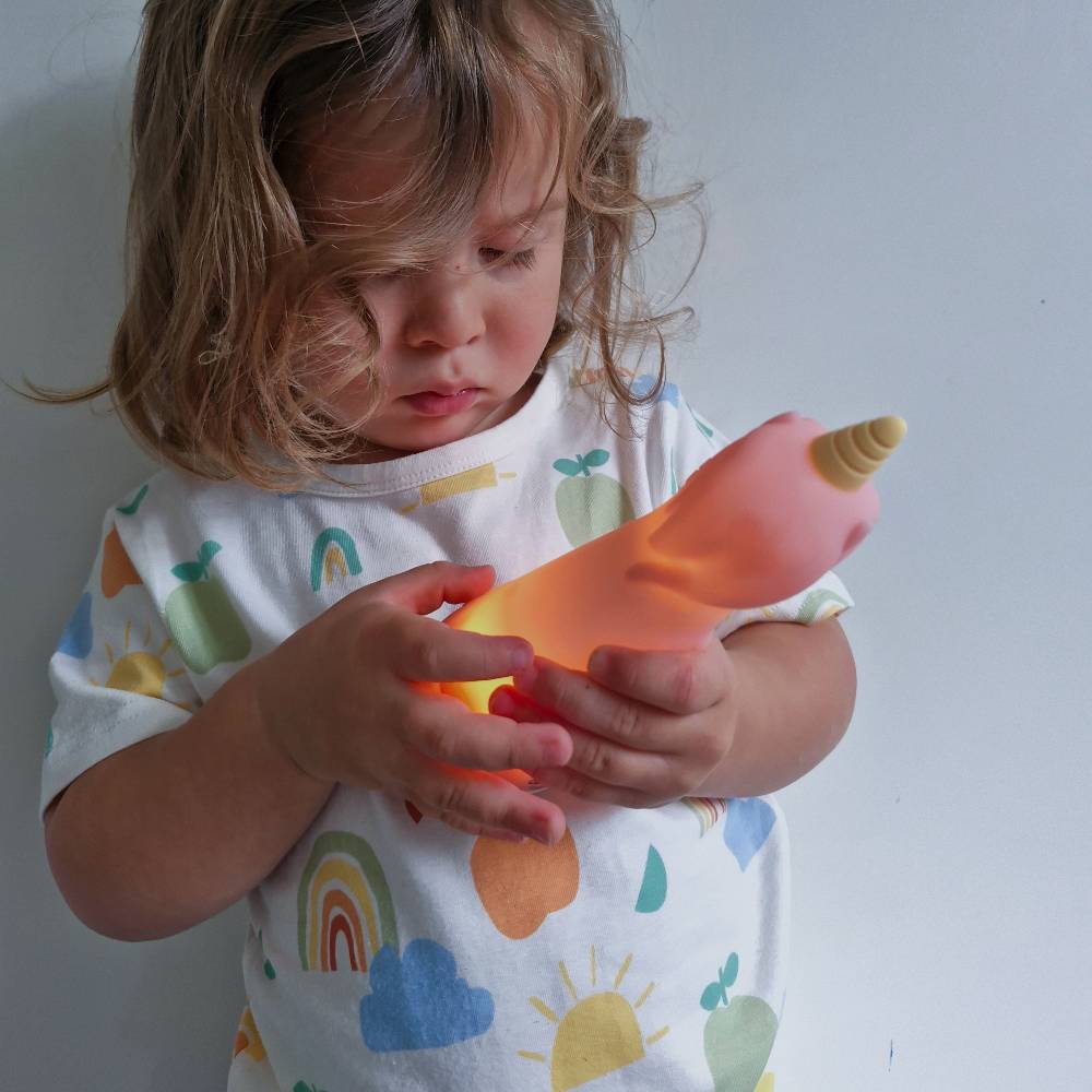 Young child wearing a colourful pyjama top holding a pink silicone unicorn torch against a white wall