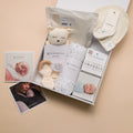 A baby gift box set from the Sleepstore, featuring a plush bear toy, swaddles, a wooden teether, a muslin blanket, and care instructions, arranged on a beige background with two printed photos.