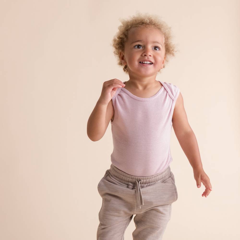 Child wearing a light pink sleeveless deluxe merino rib singletsuit and beige pants on a beige background