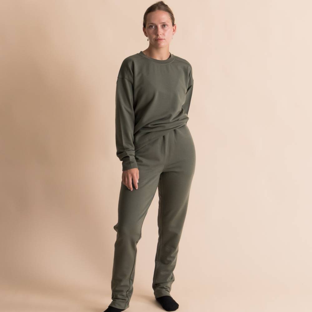 A woman modeling a matching two-piece lounge set in olive green, consisting of a long-sleeve merino fleece crewneck sweatshirt and coordinating sweatpants, against a solid beige studio background.