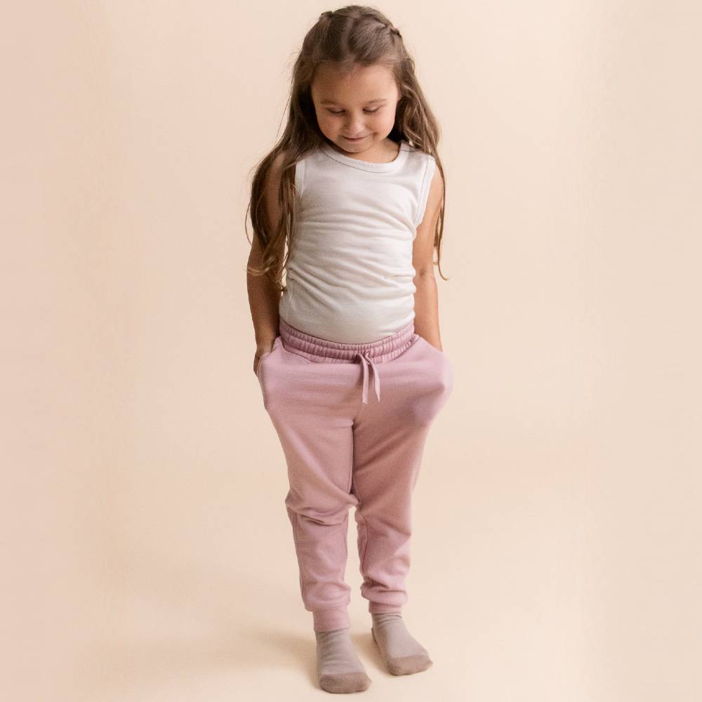 A young girl with long brown hair stands against a solid beige background, wearing dusty pink merino fleece sweatpants, a white sleeveless top, and light gray socks.