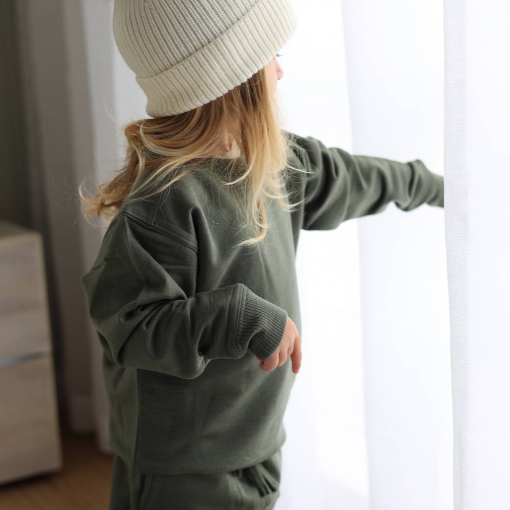 A child wearing a merino tencel/fleece olive green sweatshirt and matching pants, paired with a cream ribbed beanie, standing by a window with white curtains.
