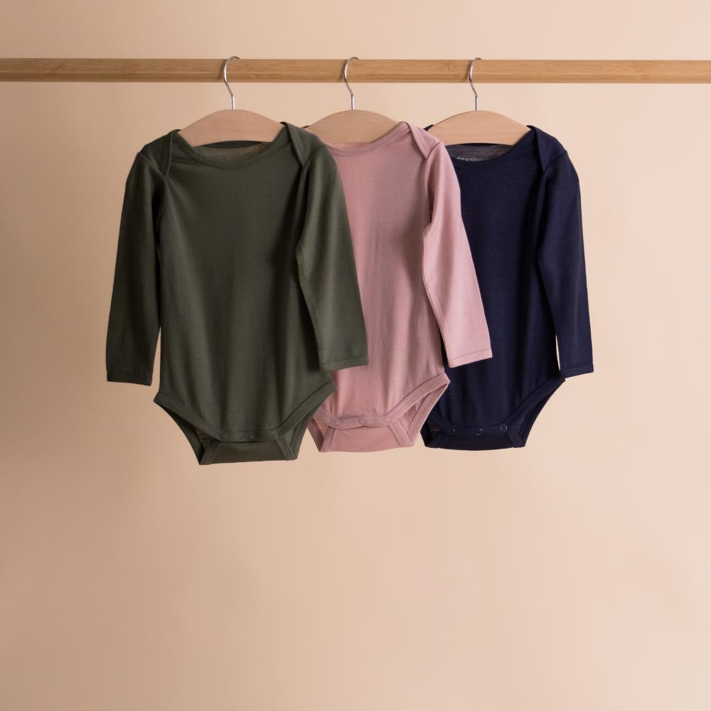 Three children's bodysuits in green, pink, and navy hanging on a rack against a beige background.