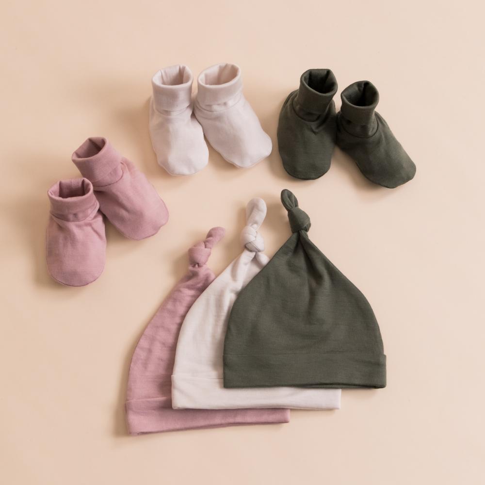 Set of Merino Tencel baby shoes and hats in pink, white, and green on a beige background
