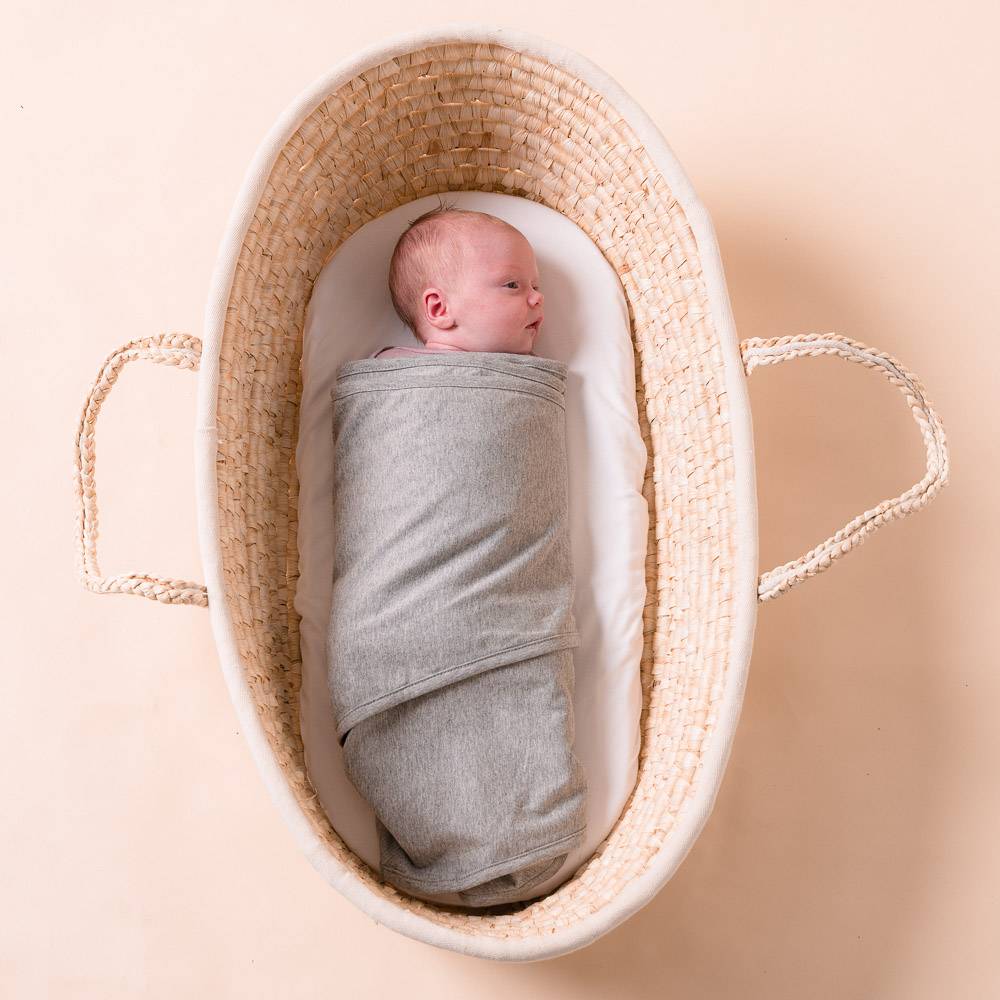 Newborn baby swaddled in a grey miracle blanket lying in a woven basket on a beige background