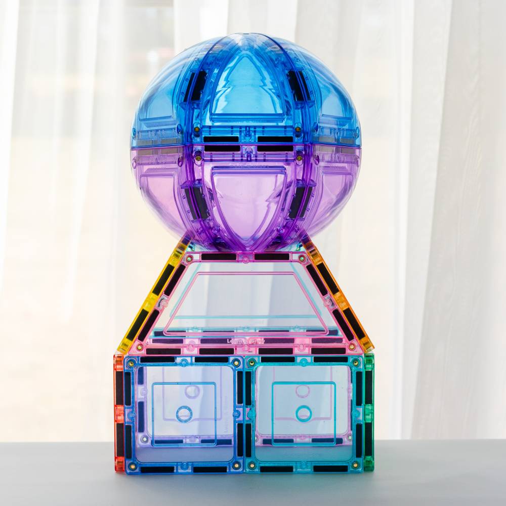 A colorful transparent magnetic building toy set arranged into a decorative structure with a translucent blue-purple spherical top, a pink and yellow triangular frame, and two small rectangular base compartments on a light surface against a soft white curtain background.