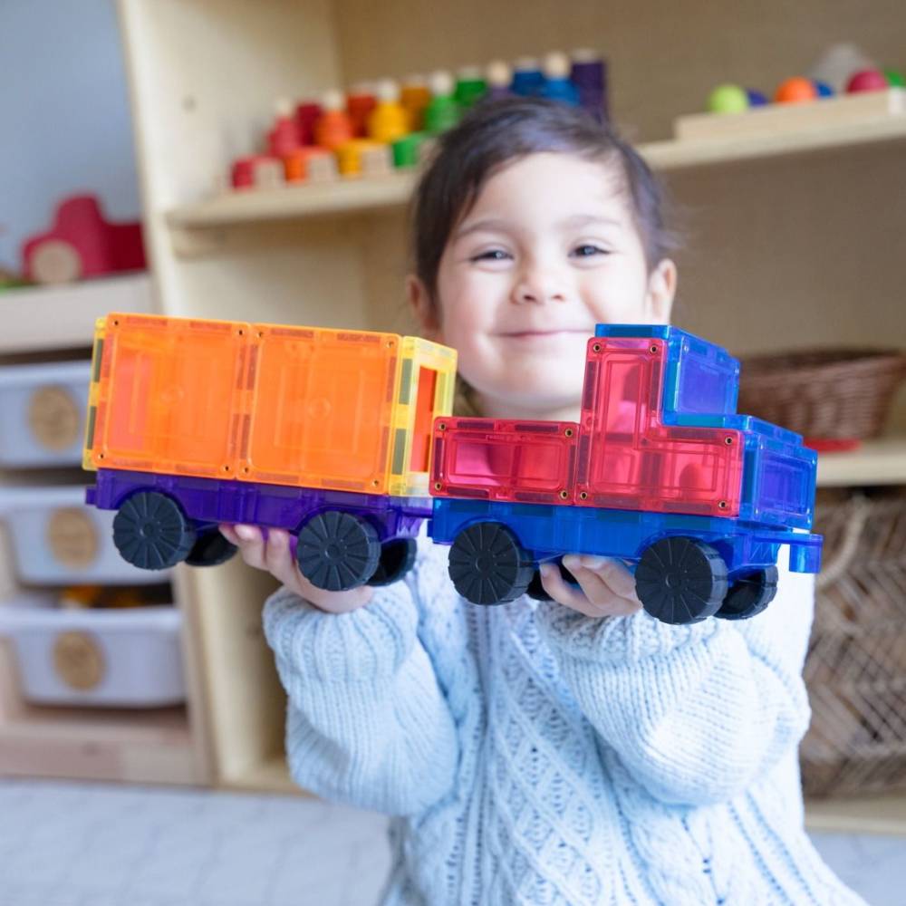 A young child smiling while holding two colorful magnetic building toy trucks made of translucent plastic tiles in a bright indoor playroom.