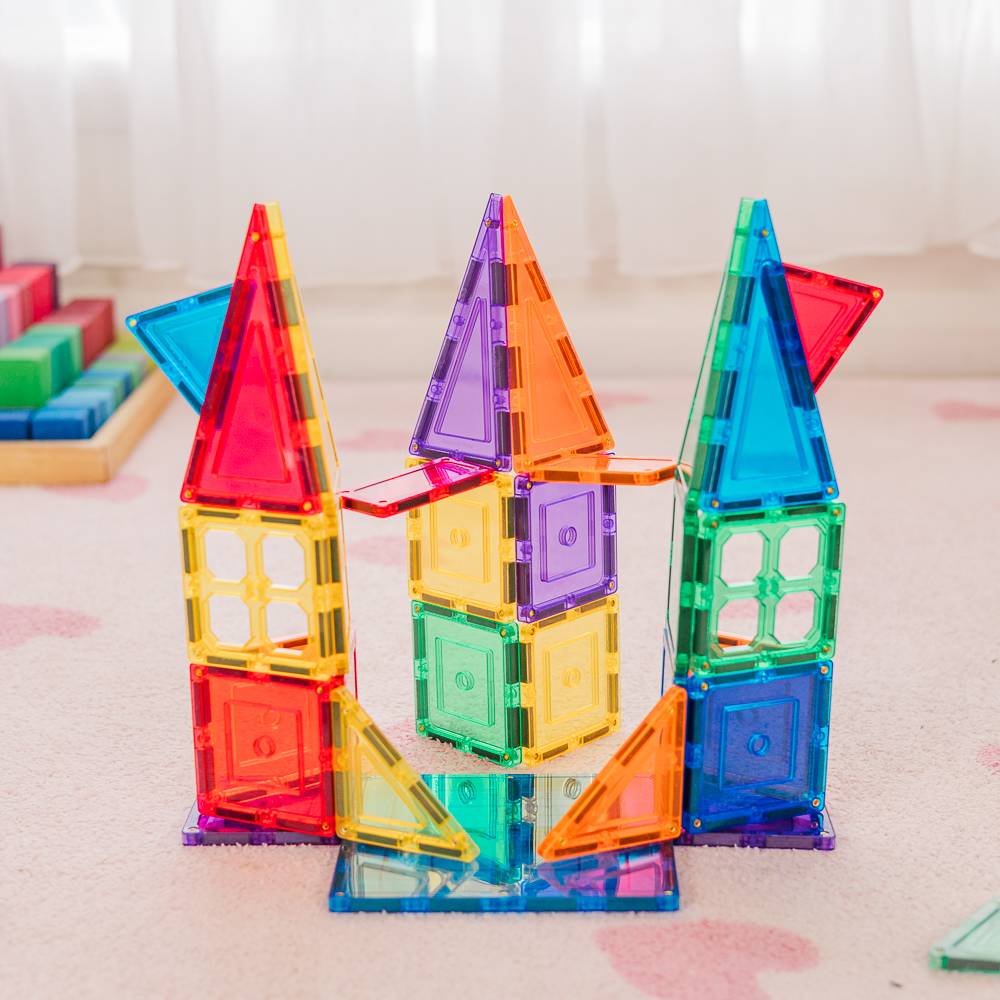 Three colorful translucent magnetic building towers made of square and triangular tiles, standing on a light pink carpet in a softly lit indoor setting.