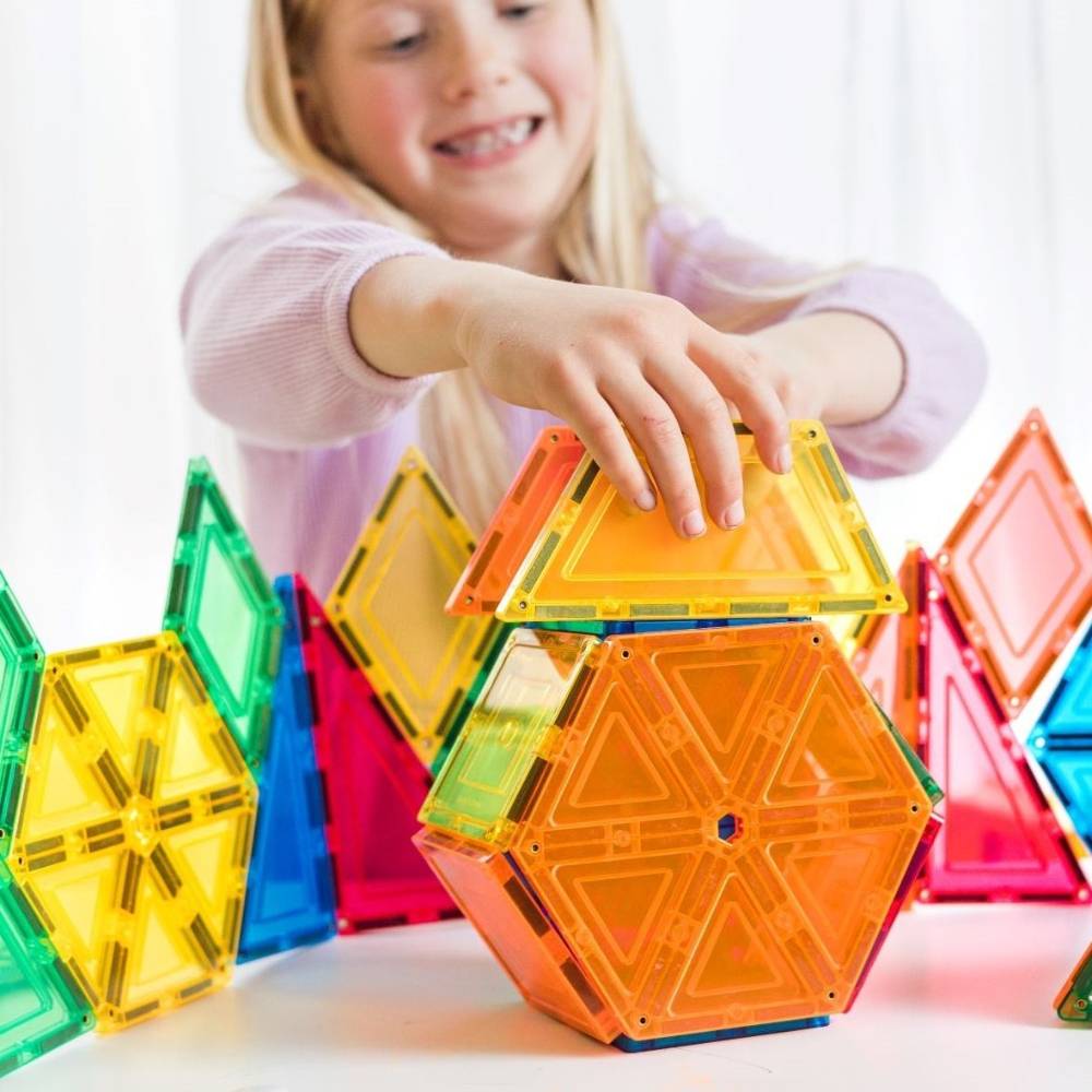 A young girl smiling and building with translucent magnetic tiles, including a prominent orange hexagonal piece and other colorful pieces on a white surface.