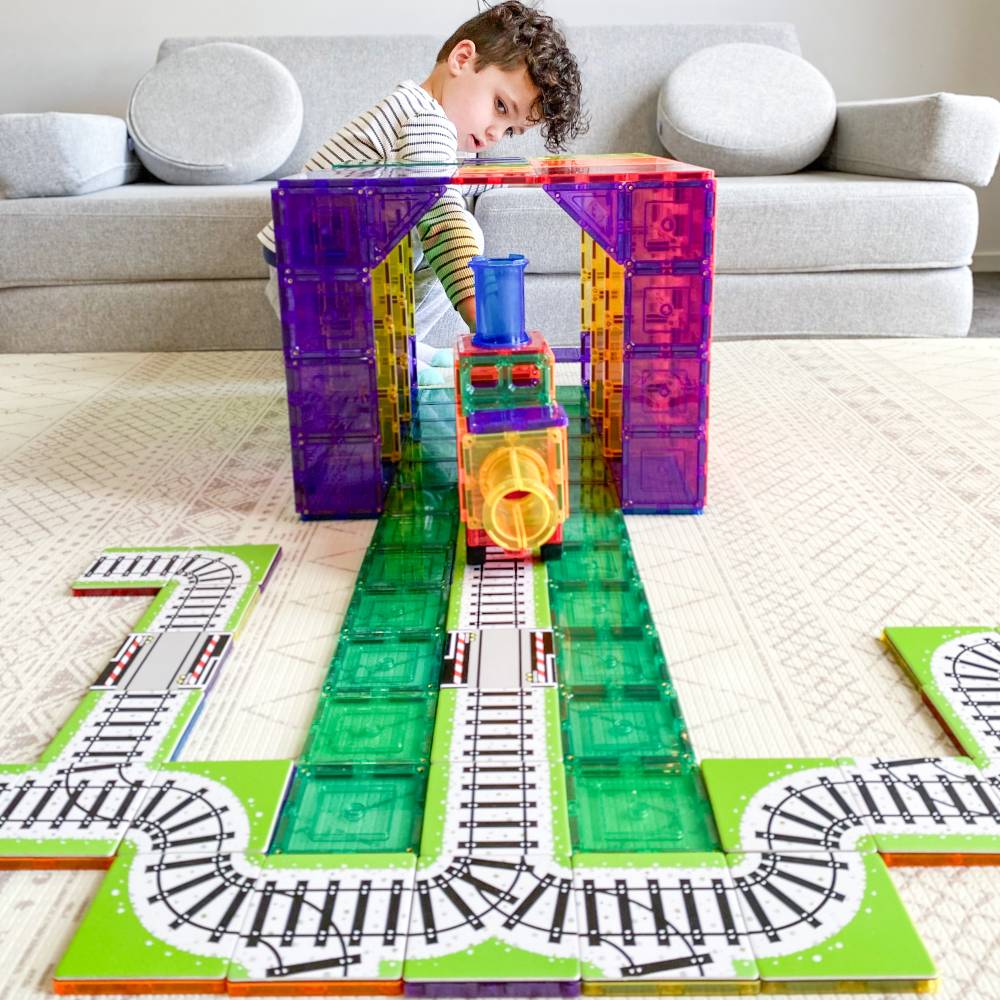 A child is building a colorful magnetic train track set on a light-colored carpet in front of a gray sofa. The set includes a purple arch, a green track bed, and a train engine with a yellow front and blue smokestack.