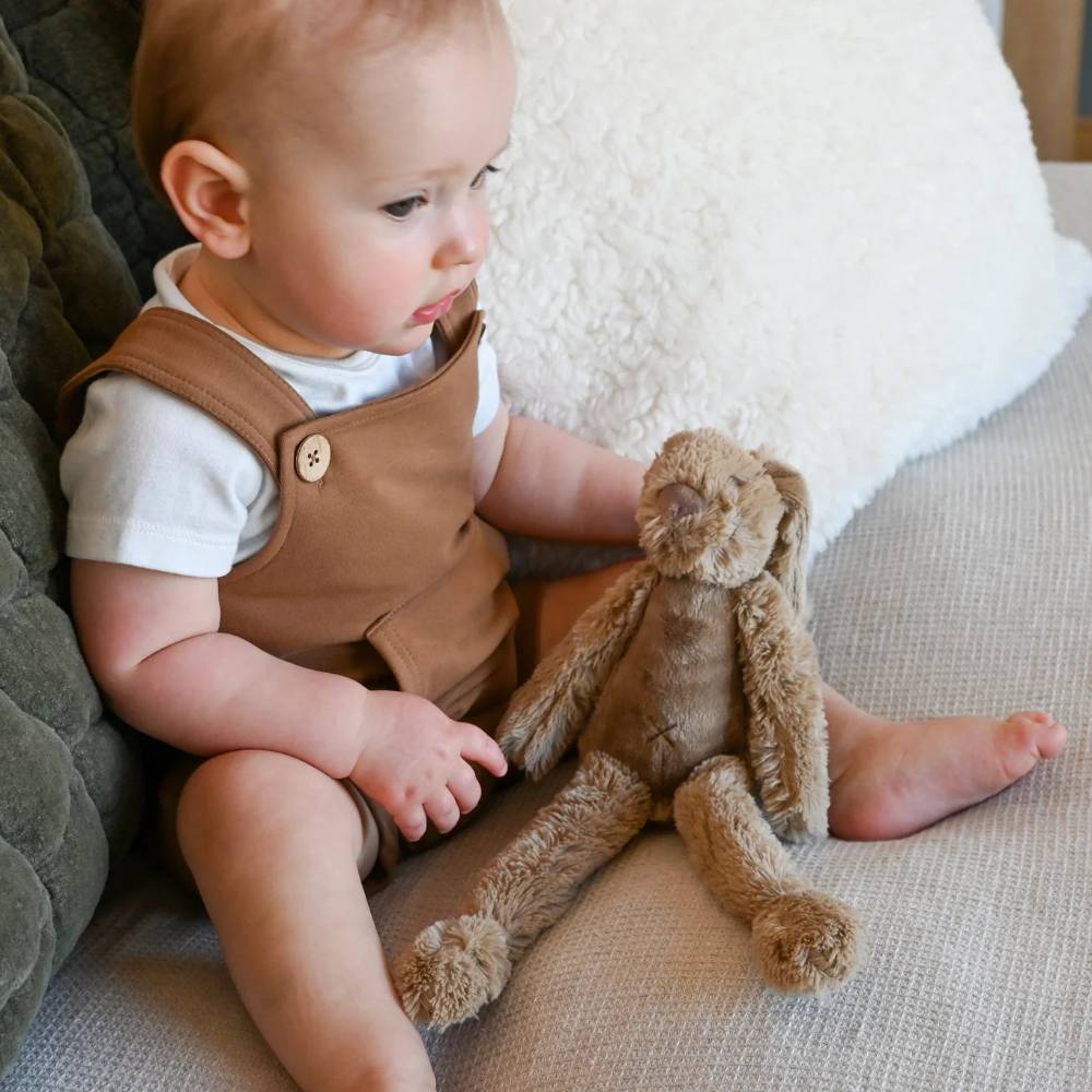 A baby sitting on a textured beige sofa, wearing a brown sleeveless romper over a white t-shirt and holding a brown plush bunny toy.
