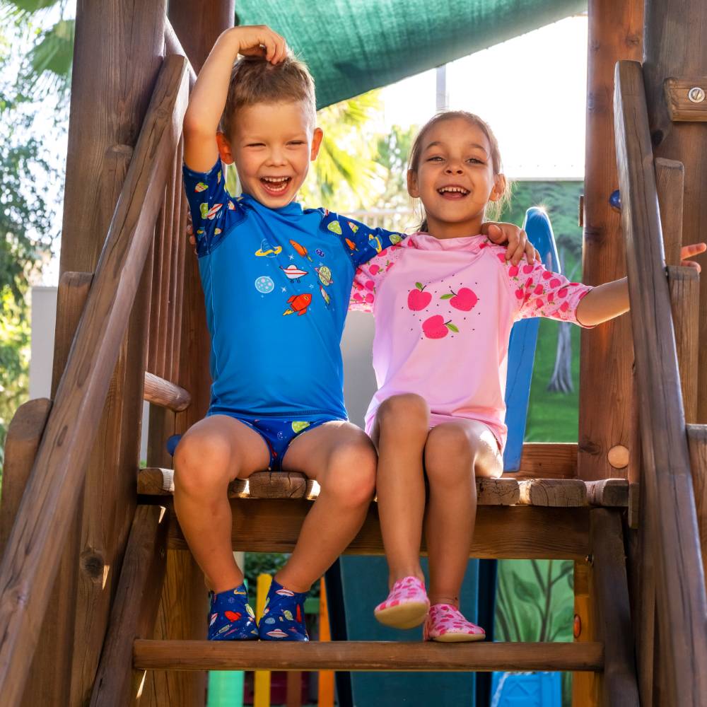 Two children sitting on a wooden playground slide wearing matching swim shirts and shorts sets. The boy wears a blue space-themed swim set with yellow and blue accents, and the girl wears a pink swim set with red and yellow fruit graphics. They wear matching slipfree water shoes
