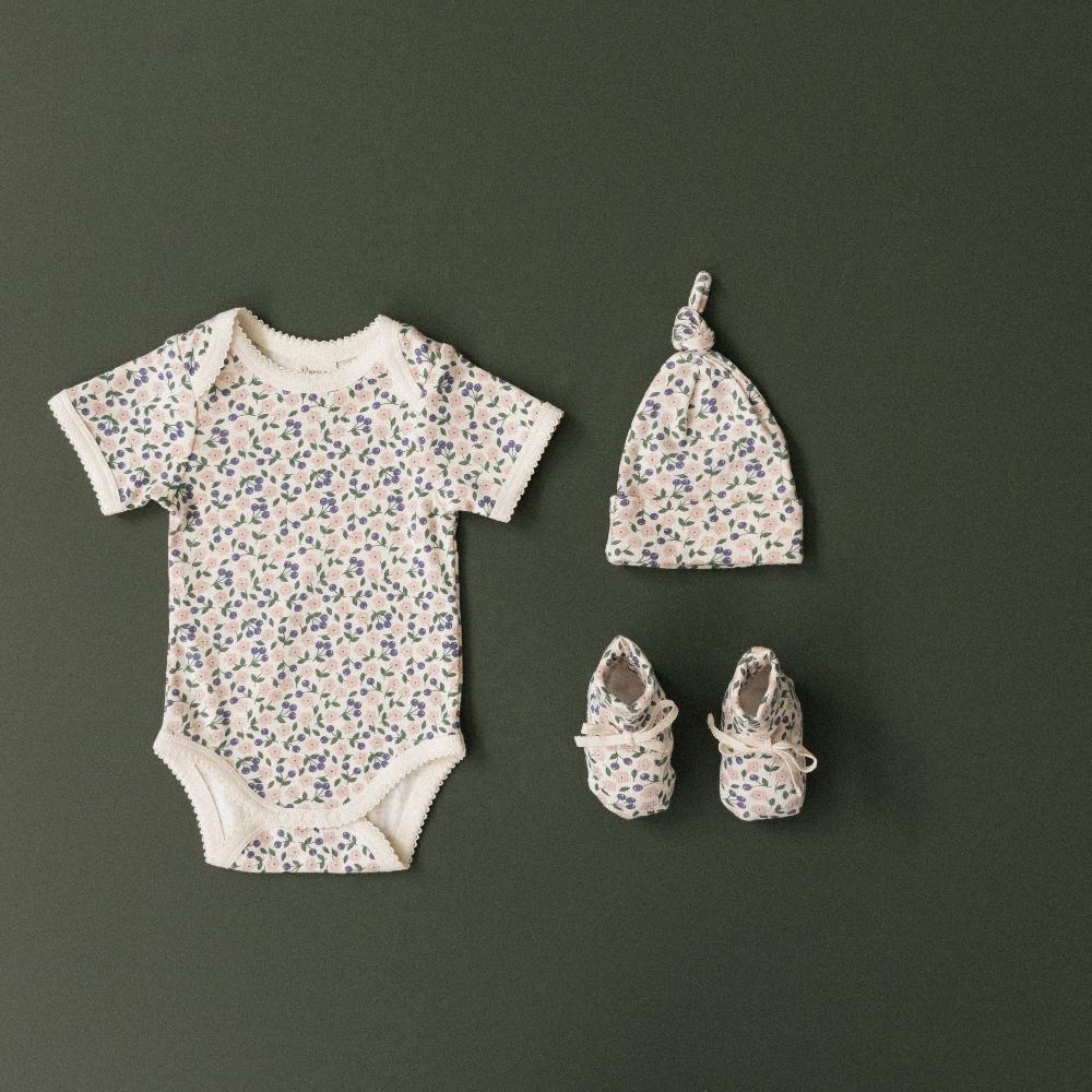 Baby clothing set with cream and purple/pink floral-patterned bodysuit, hat, and shoes on a dark green background