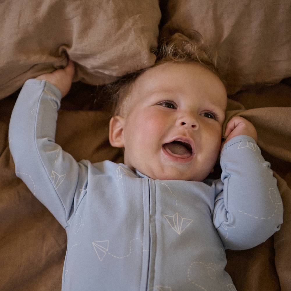 Baby in a light blue organic cotton onesie with white paper plane pattern lying on brown bedding