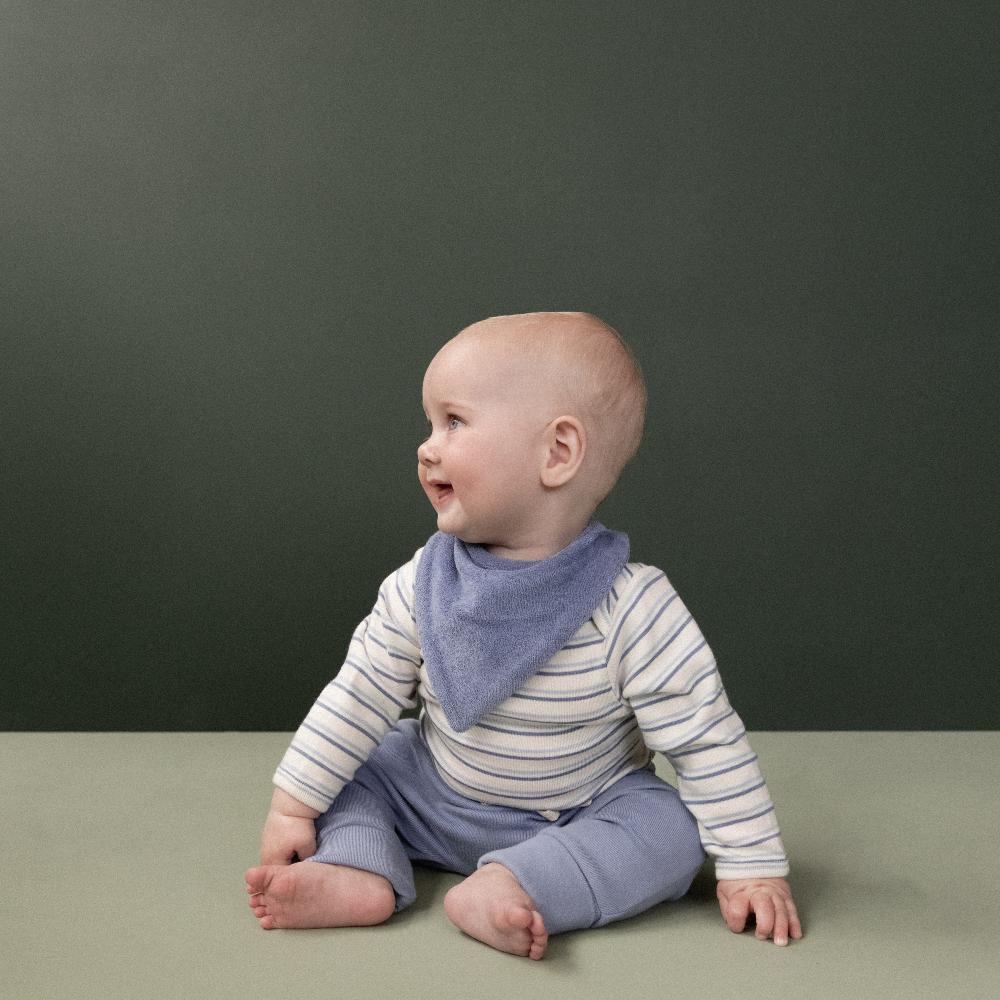 Baby sitting on a green surface with a dark gray background wearing a striped bodysuit and light blue bants to match 
