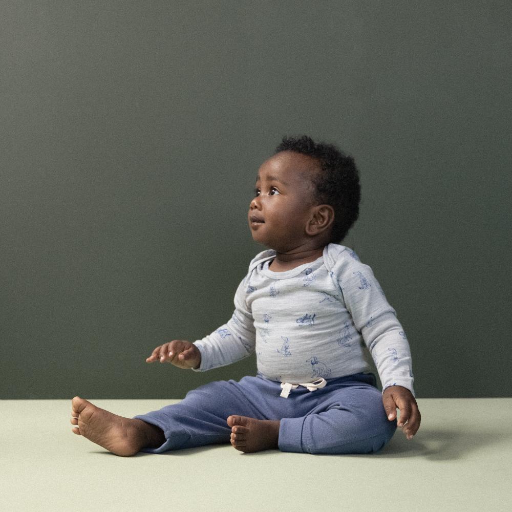 Baby sitting on a light surface with a dark gray background wearing blue baby pants and grey merino bodysuit 