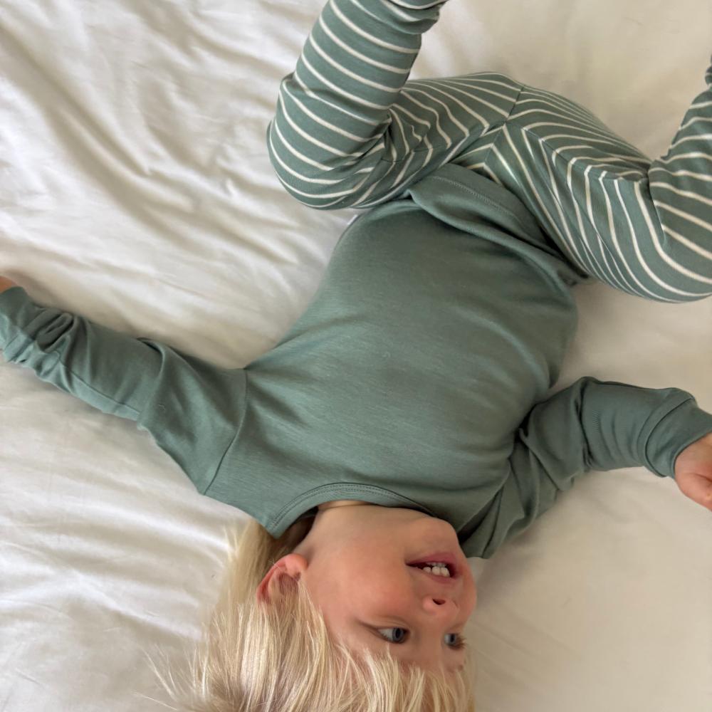 A child lying on white bedding, wearing a sage green jersey merino long-sleeve top and matching striped pajama pants in green and white.
