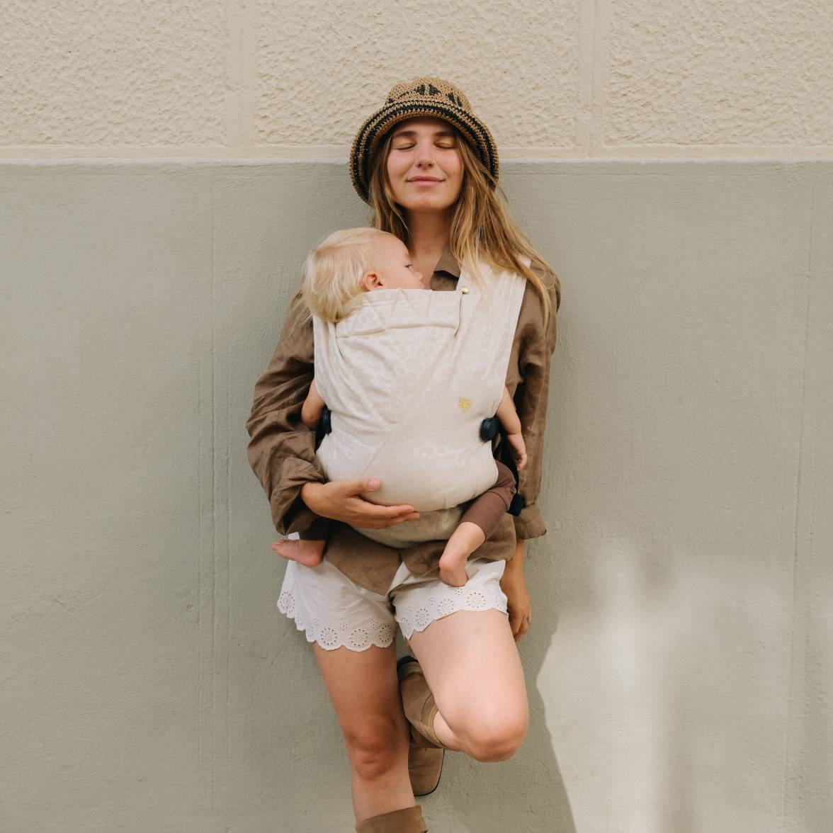 A woman resting on a wall wearing her baby in a beige leopard print baby carrier on the front facing in. 