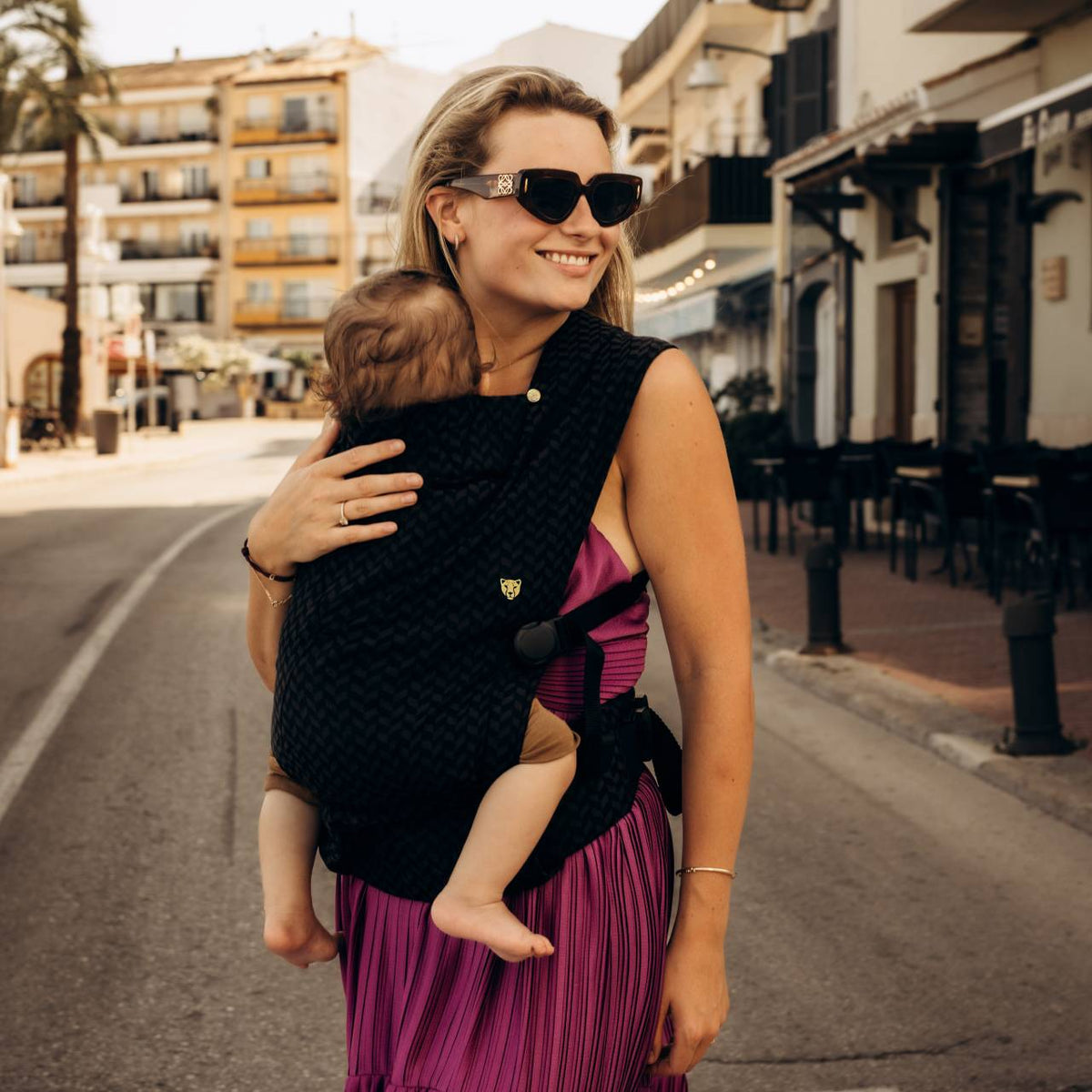 A woman walking down the street, carrying her baby in a black baby carrier on the front facing in. 