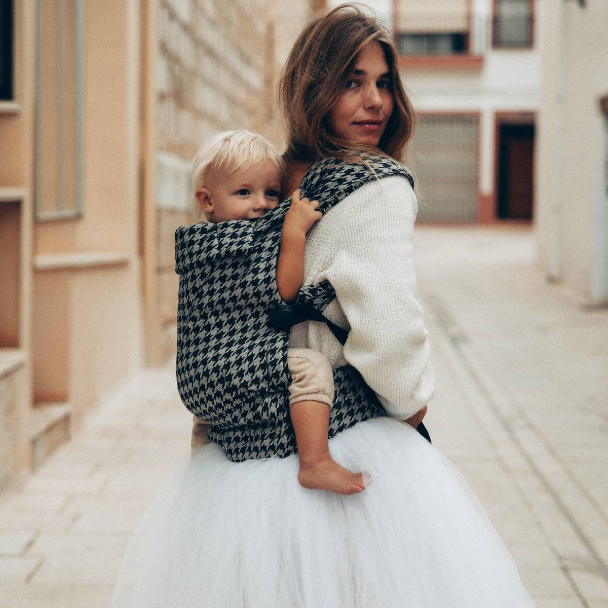 A woman wearing her toddler in a black and grey houndstooth patterned baby carrier on her back. 
