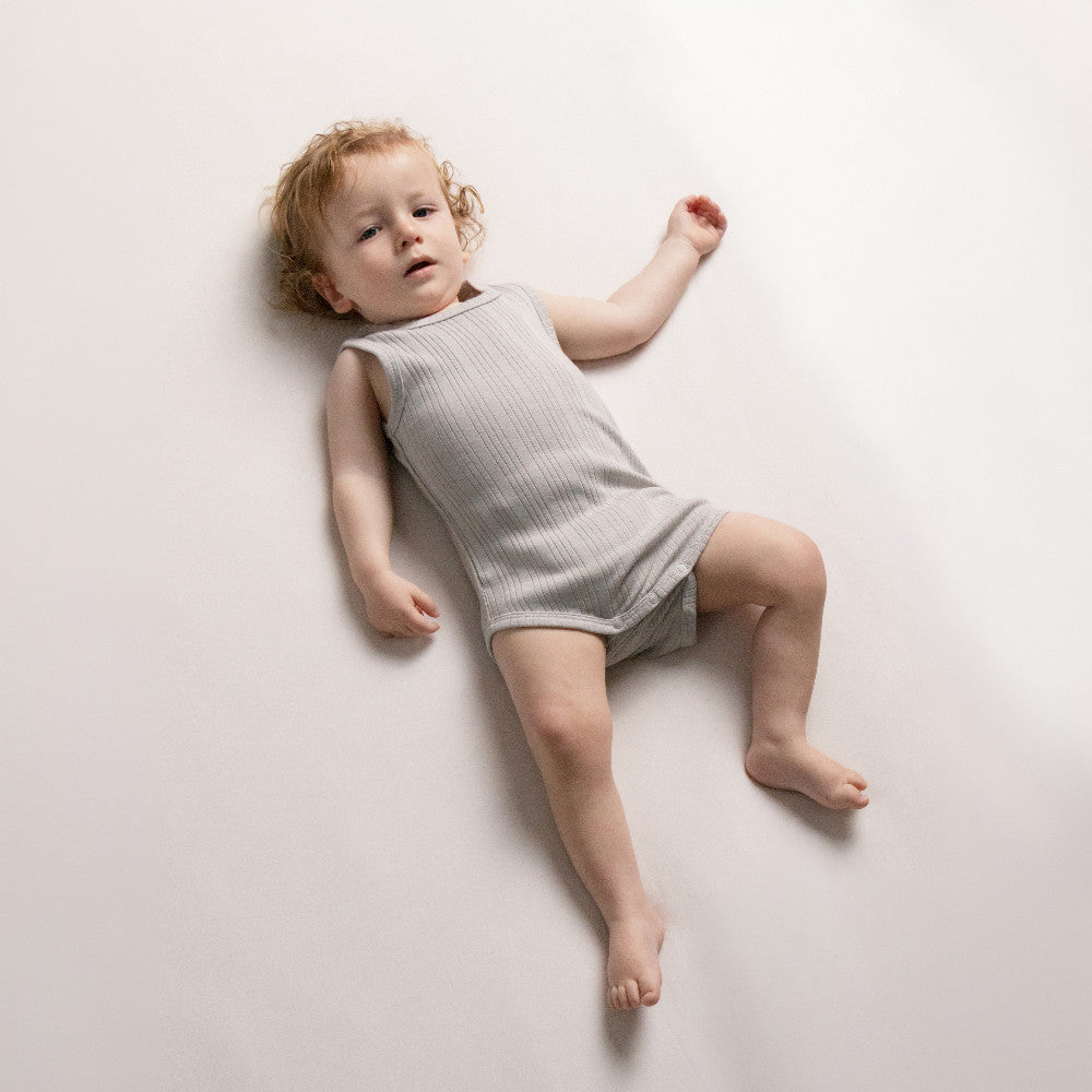 Baby lying on a white surface wearing a pebble marl Woolbabe Merino Cotton Rib Singletsuit. 