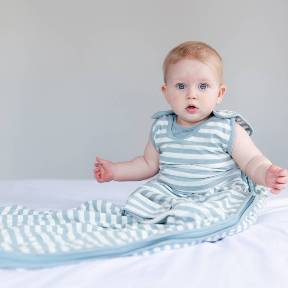 Baby in a blue and white striped woolbabe sleeping bag sitting on a bed.