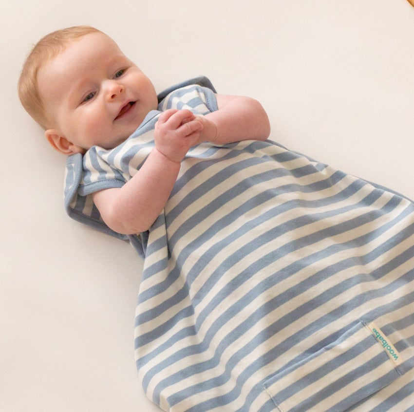 A baby lying on a cream-colored surface, wrapped in a blue and cream striped sleep sack. The brand name 'woolbabe' is visible on a small tag on the lower right of the sleep sack.
