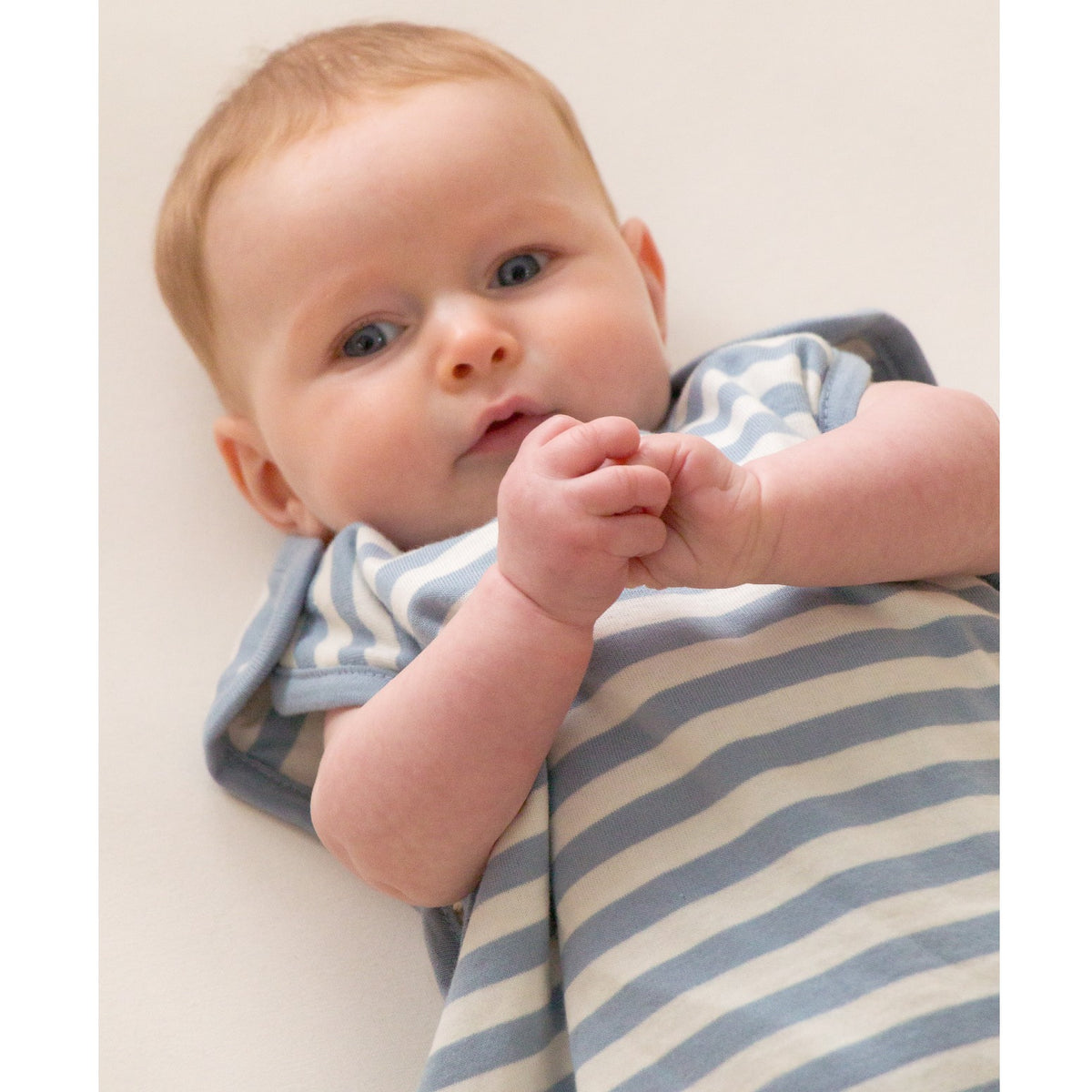 A close-up of a baby lying on their back, wearing a light blue and white striped sleeping bag with shoulder snaps 