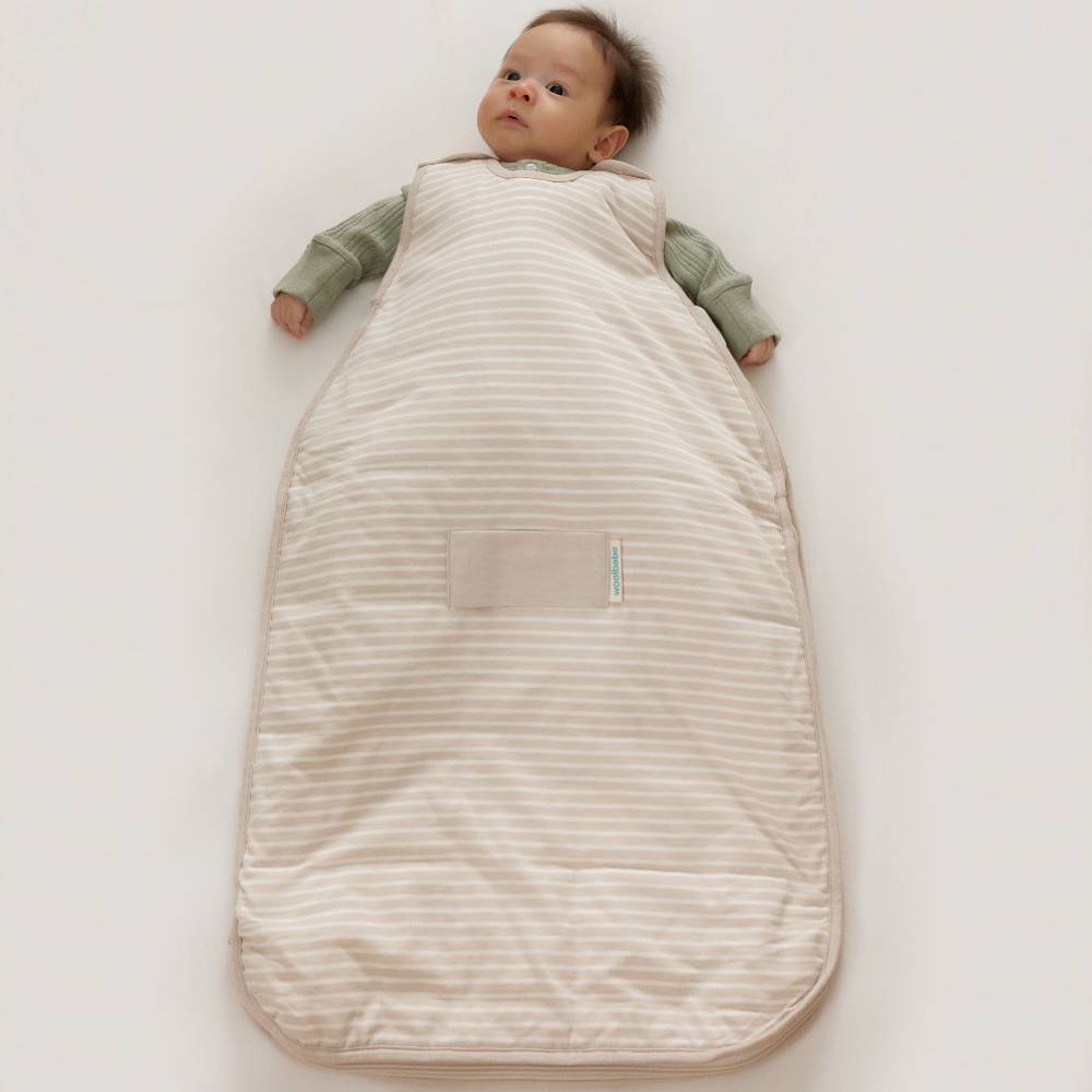 A baby lying on their back wearing a cream and beige striped sleep sack over an olive green long-sleeve shirt, photographed on a clean white background.