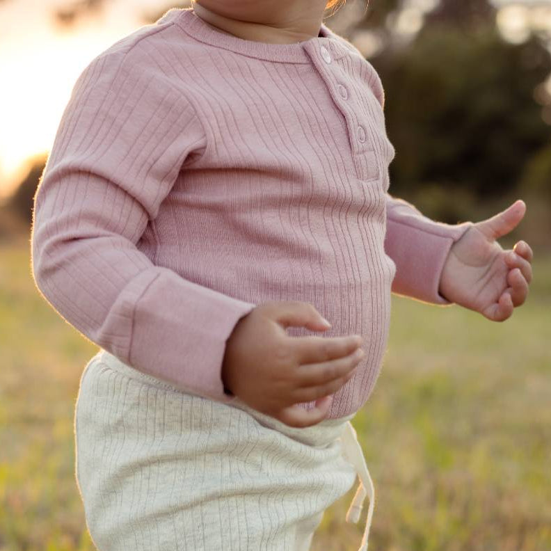 Child wearing a pink rib bodysuit and white pants standing in a grassy field.