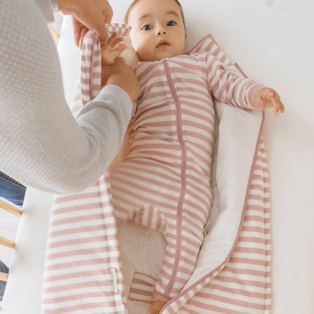 Baby in a pink and white striped outfit being held by an adult on a white blanket.