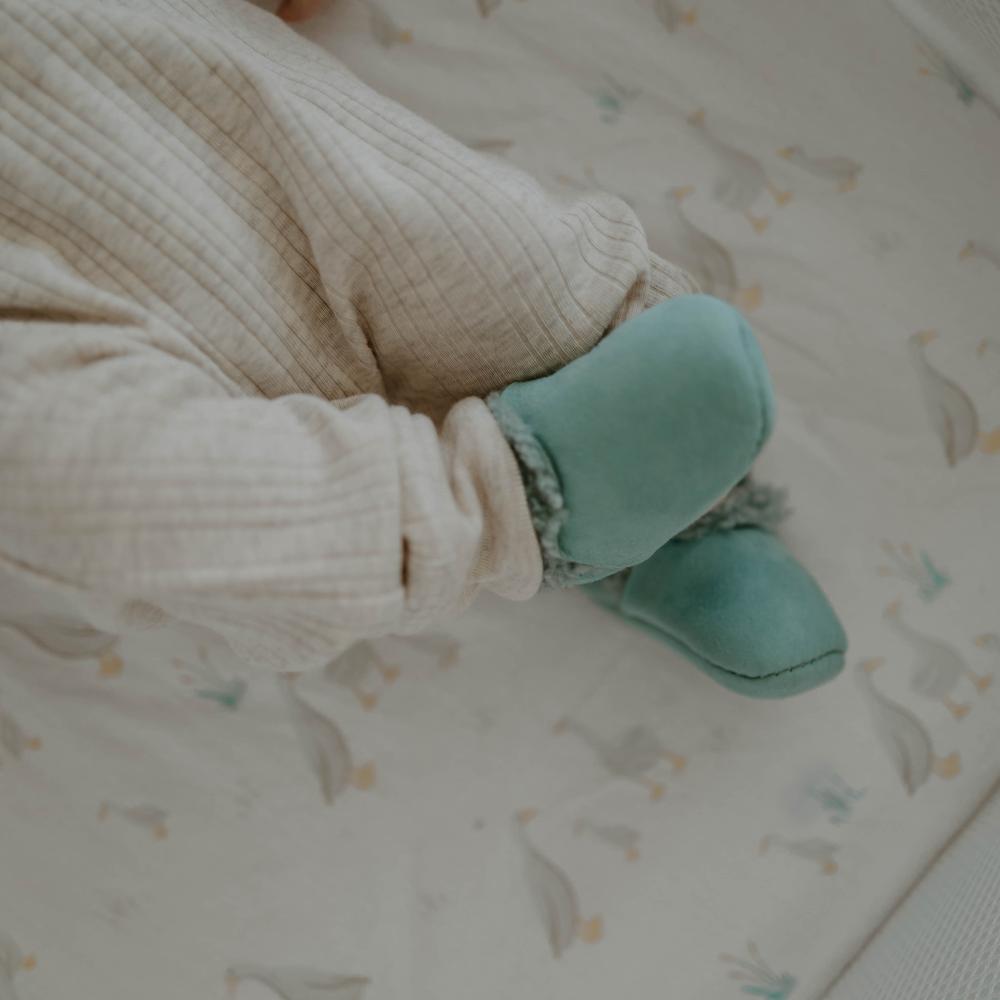 Baby wearing tide green lambkins booties on a crib with a patterned sheet