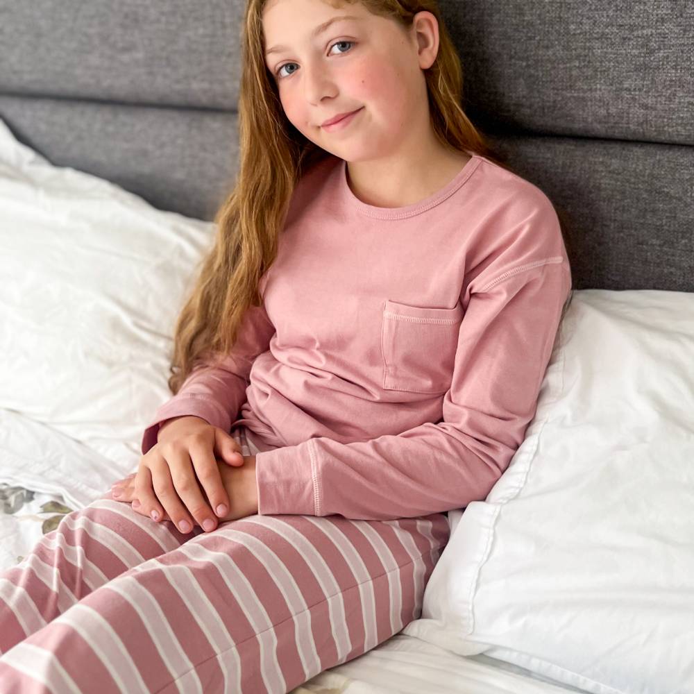 Young girl in pink pajama top and striped pants sitting on a bed.