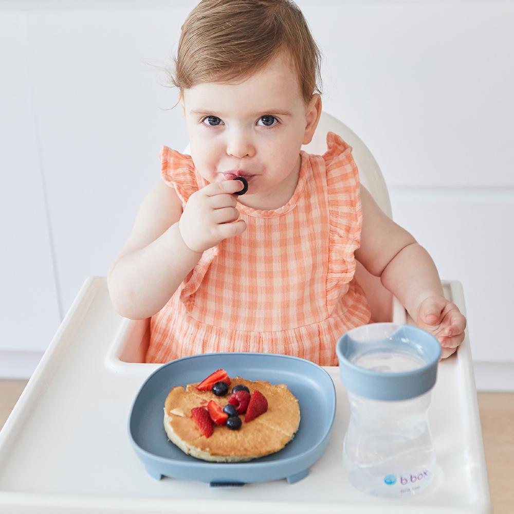 Child in a high chair eating a pancake with berries on a suction plate and a blue cup on a white background