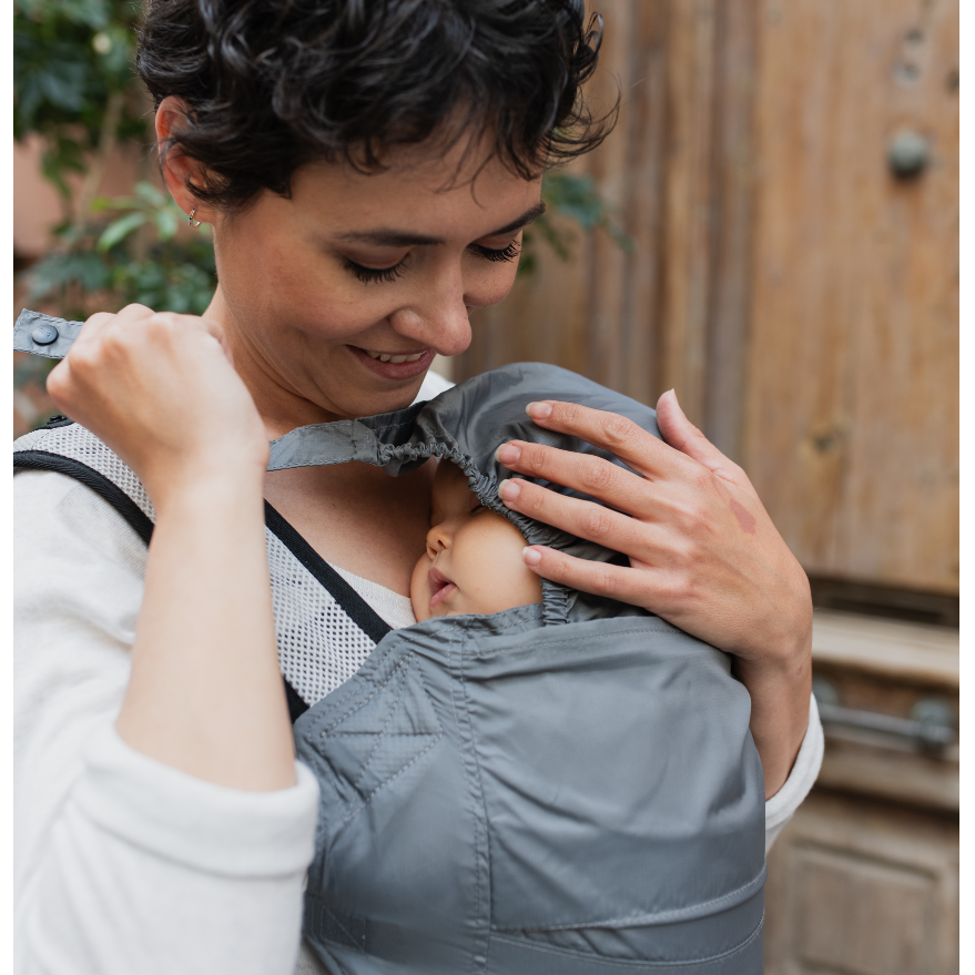 Woman holding a baby in a gray baby carrier outdoors.
