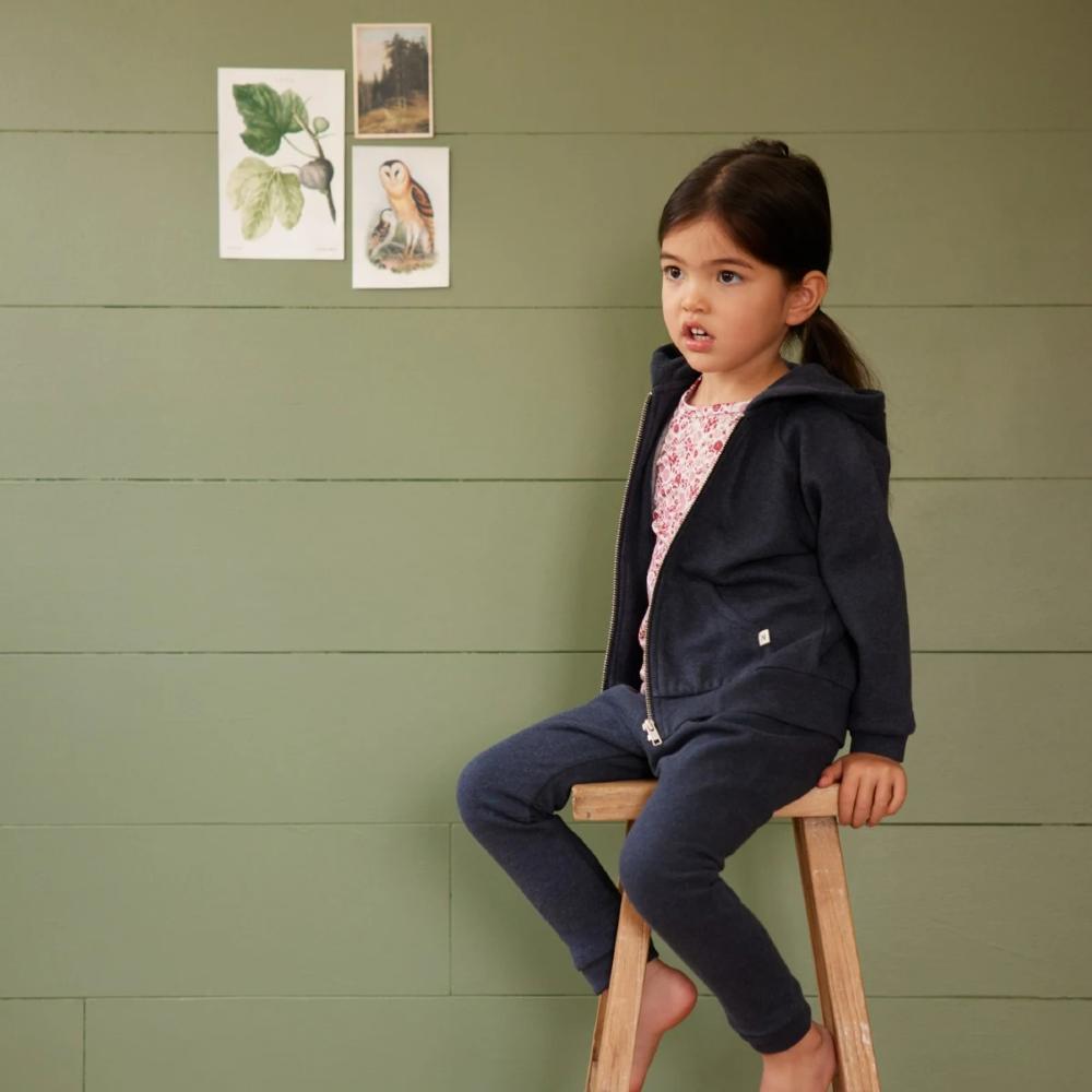 Young girl sitting on a stool against a green wall with nature-themed posters wearing a matching navy tracksuit set by NatureBaby