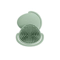 A green silicone scalp massager brush with a matching lid, shown open on a solid white background. The brand name 'shnuggle' is embossed on the lid.